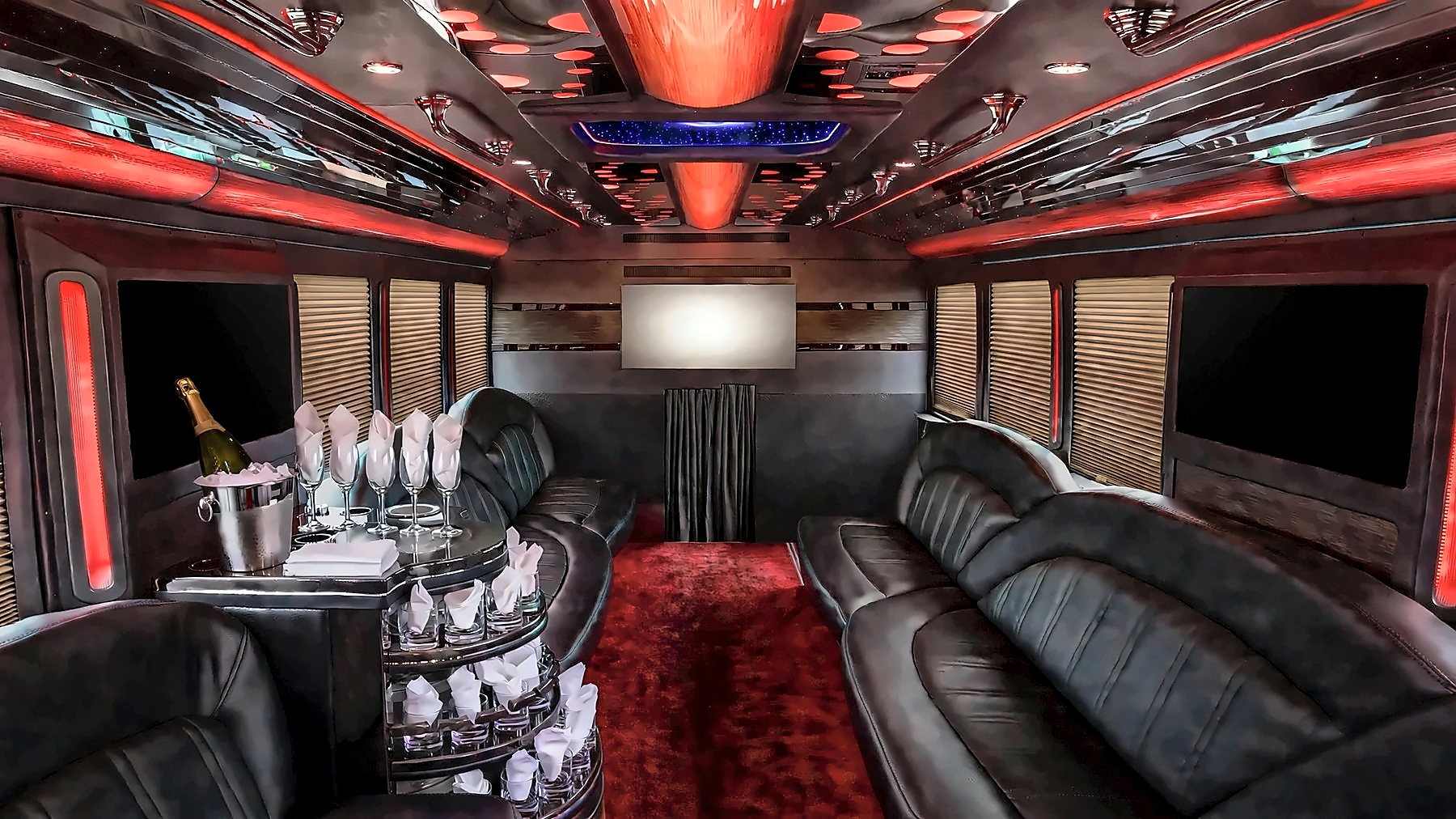 Interior of the Ford F-550 Party Bus looking toward the front, with leather bench seating, a bar with glassware, a red carpet aisle and red cabin lighting