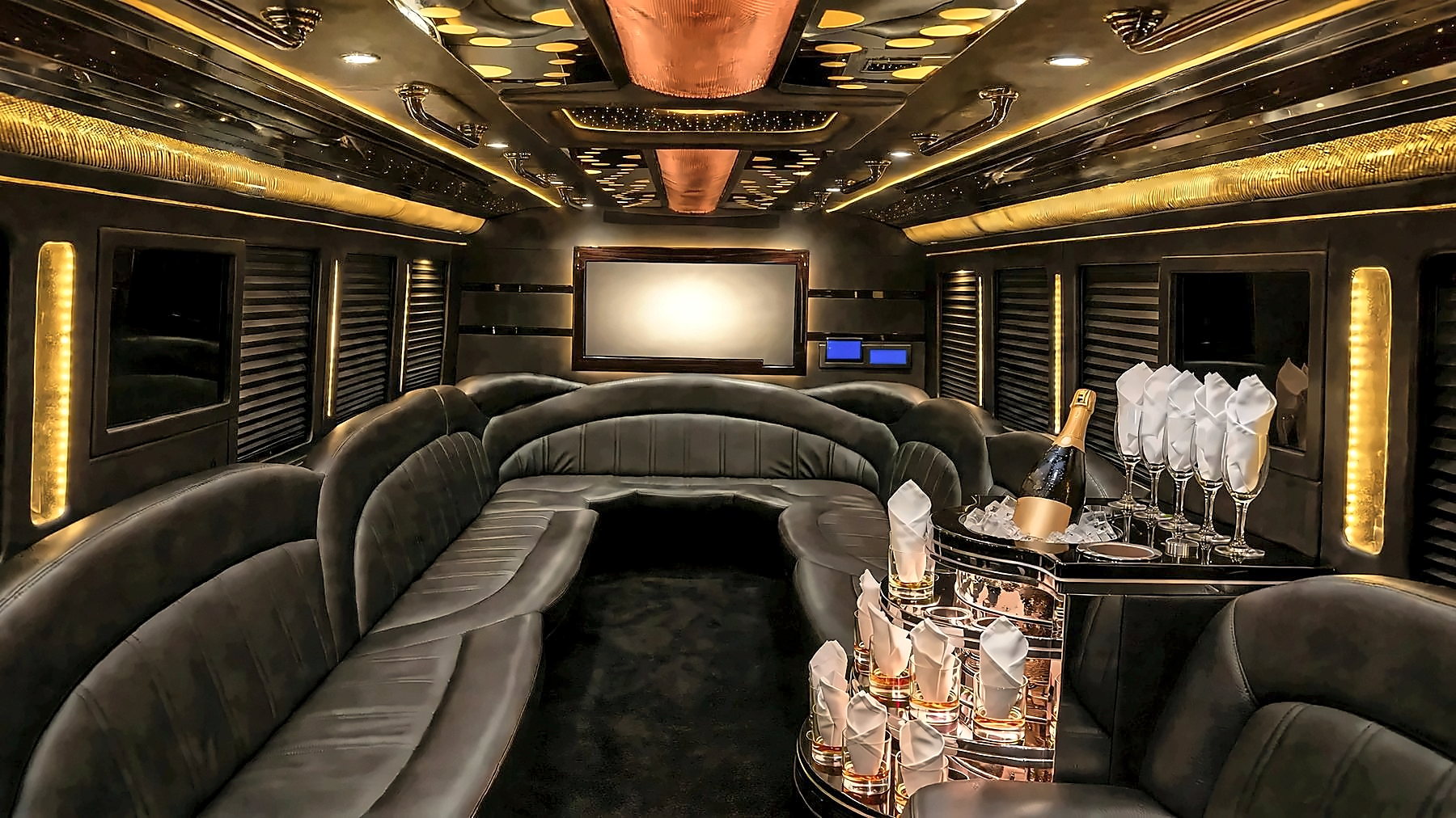 Interior of the Ford F-550 Party Bus with gold cabin lighting, wraparound leather lounge seating at the back, a starlight and mirrored ceiling, a screen and a bar with glassware