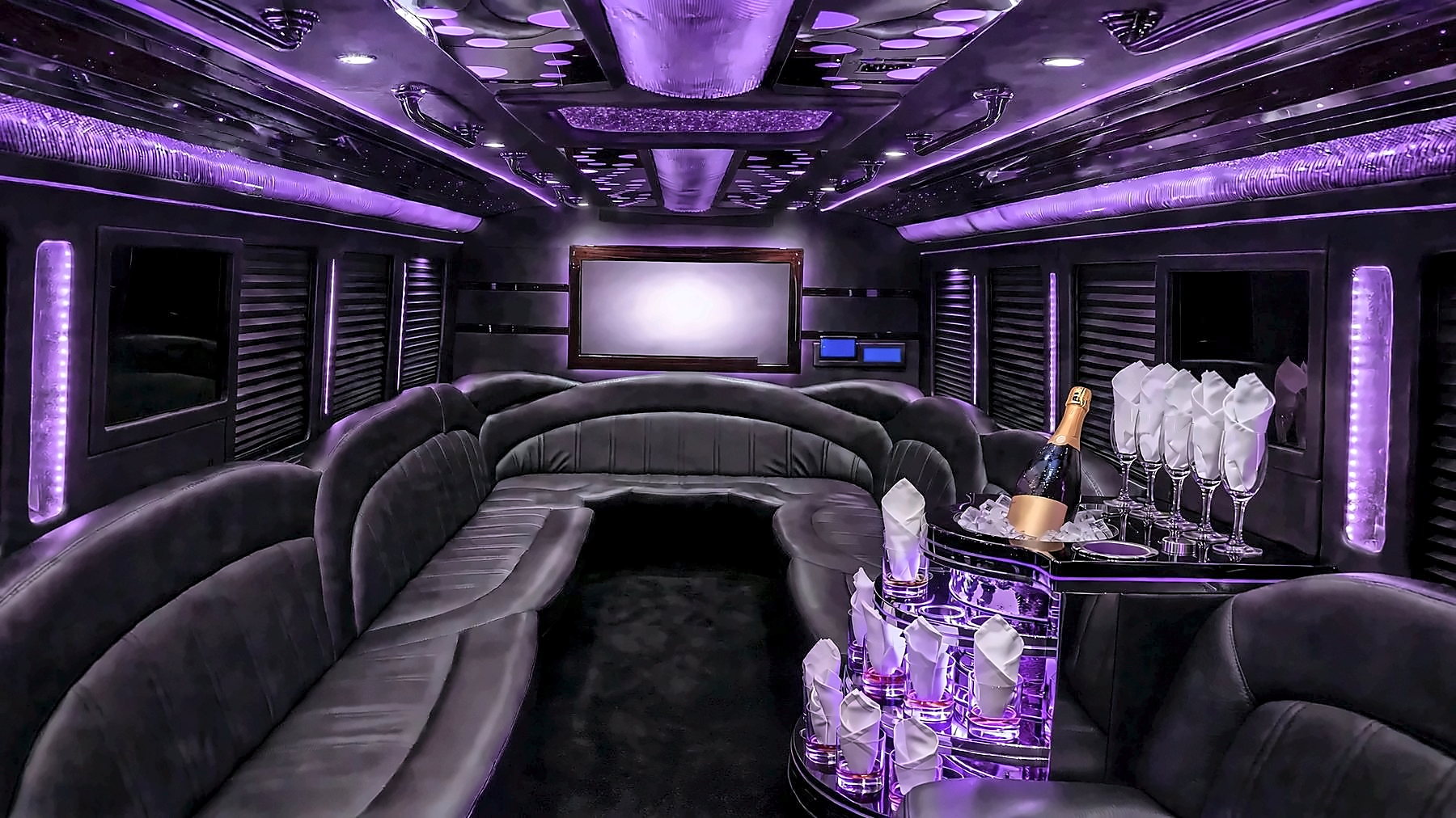 Interior of the Ford F-550 Party Bus with purple cabin lighting, wraparound leather lounge seating at the back, a starlight and mirrored ceiling, a screen and a bar with glassware