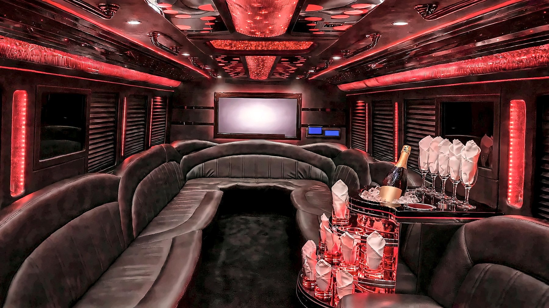 Interior of the Ford F-550 Party Bus with red cabin lighting, wraparound leather lounge seating at the back, a starlight and mirrored ceiling, a screen and a bar with glassware